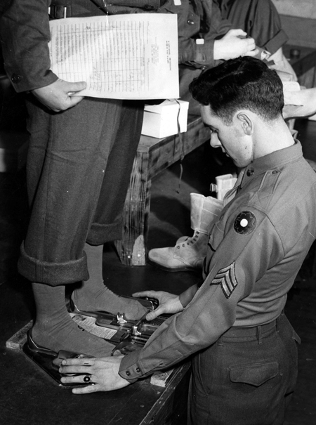 A young Army sergeant wearing his insignia and a 9th Infantry Division patch on his left arm measures another soldier’s feet, using a double Brannock device to measure both feet at once.