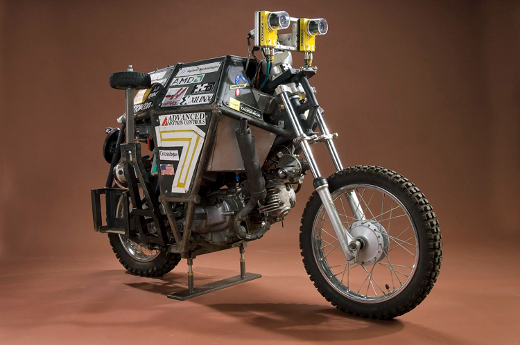 Side view of the Ghostrider autonomous motorcycle. There is a box covering the seat area and it is covered in stickers, including Agilent Technologies and an American flag. Two cameras are located where the handlebars would normally be.