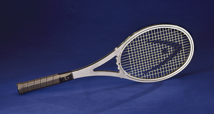 A silver metal tennis racket with a brown leather-wrapped handle sits on a cobalt blue background. The racket bears the logo of Head tennis rackets on the head and shaft, and the rim reads, “Arthur Ashe Competition.”