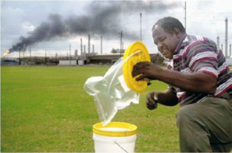 A man inserting a plastic collection bag into a 5-gallon plastic bucket, with a refinery belching smoke in the background.
