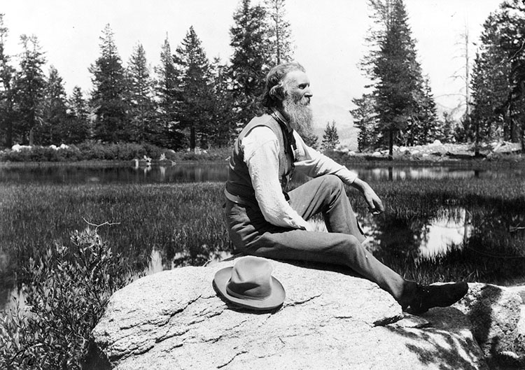 Profile view of John Muir sitting on a rock on a lake’s edge