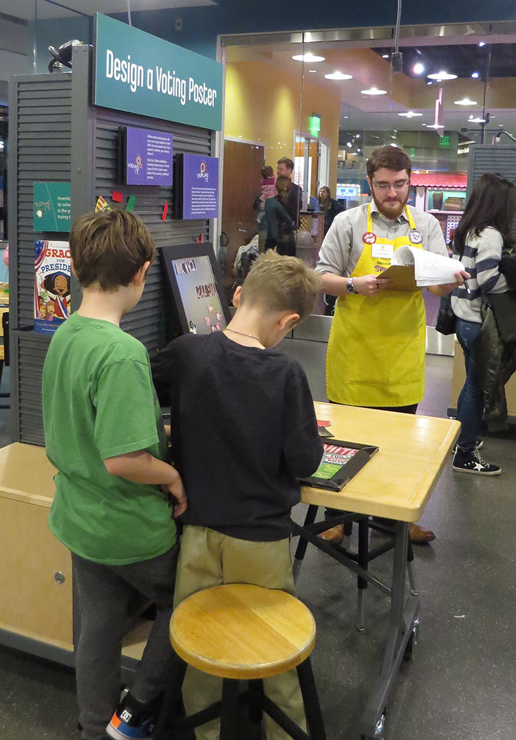 Two young boys with their backs to the camera work at the “Design a Voting Poster” activity table in SparkLab. Lead facilitator Zach Etsch, wearing a yellow SparkLab apron, holds a clipboard and papers used to evaluate the young boys’ experience.