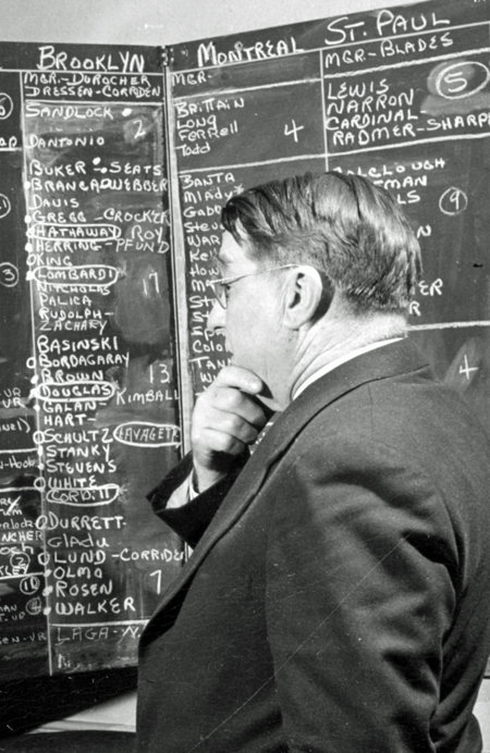 Branch Rickey holding his chin and looking at a blackboard with lists of players’ names