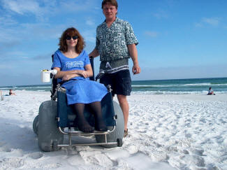 nventor Michael Deming with his wife Karen on a beach. Karen sits in a wheelchair with all-terrain wheels, specifically invented by Deming to be used on the beach.