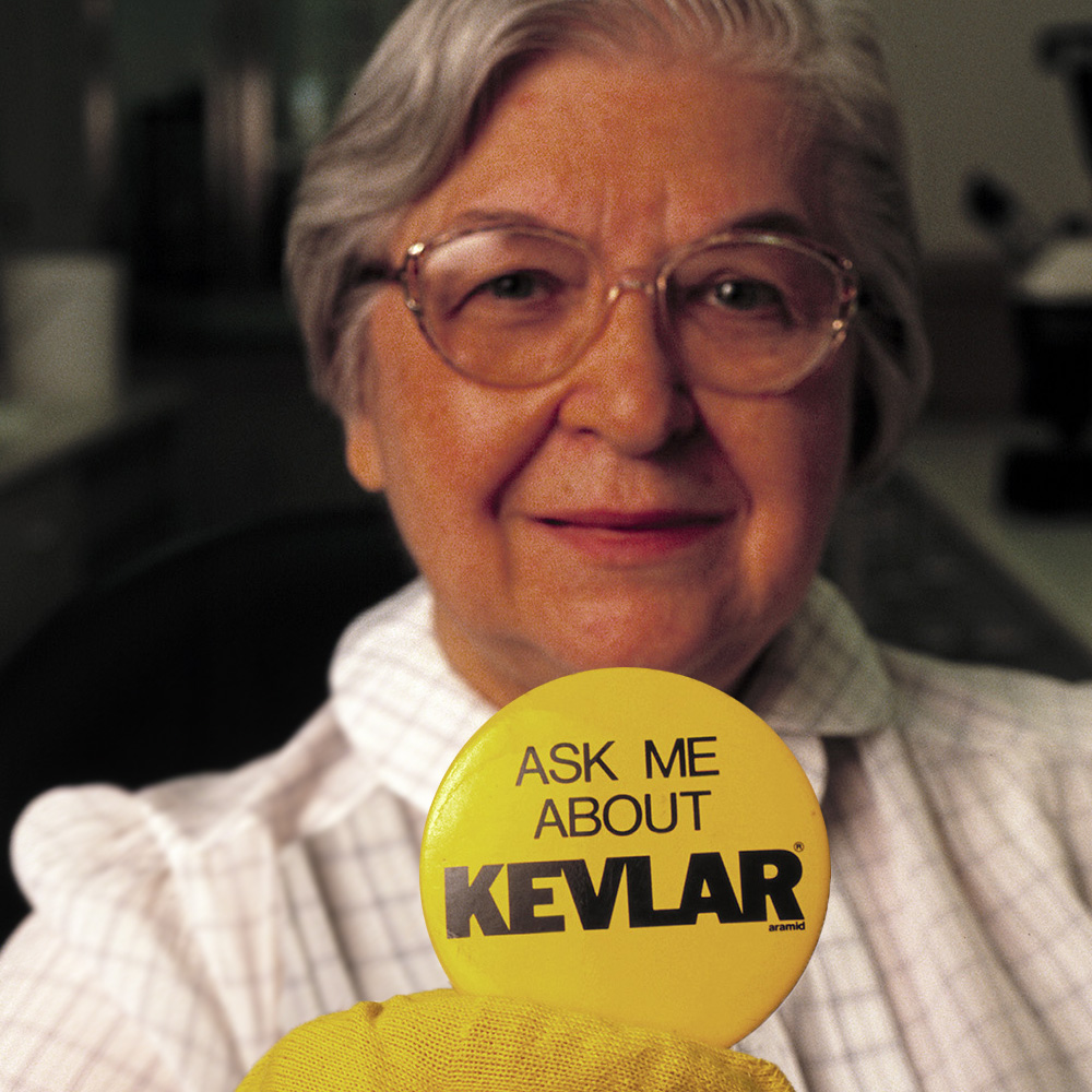Stephanie Kwolek, approximately 70 years old, holding up a button that reads Ask Me About Kevlar.