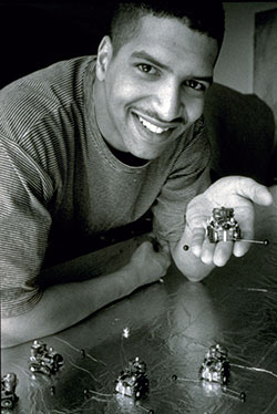 A young, adult Black man, smiling at the camera and holding a small robot with 4 wheels and 2 antennae in the palm of his hand.