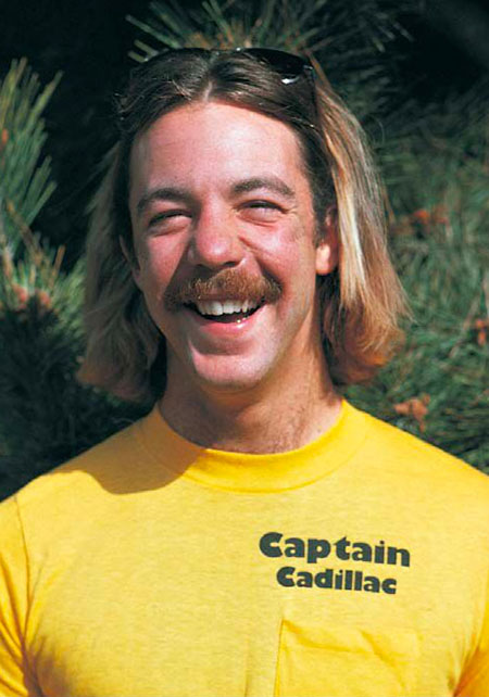 A White man in his 20s with shoulder length sandy blonde hair, smiling at the camera. A pair of sunglasses is perched atop his head and he is wearing a yellow tee shirt that reads “Captain Cadillac” a above the pocket.