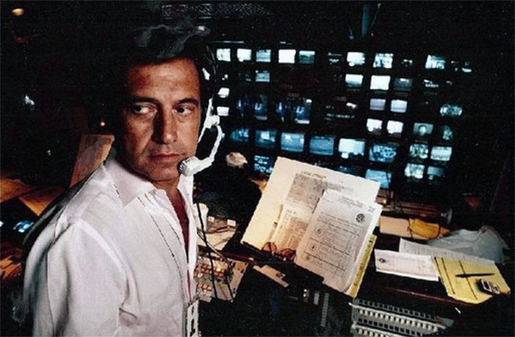 Tony Verna wearing a headset, looking over his shoulder, with papers in front of him and a bank of tv monitors in the background.