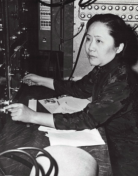 Chien-Shiung Wu in profile, sitting at a desk with a panel of knobs and dials in front of her.