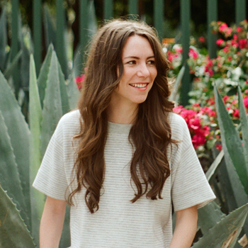 Three-quarters candid photo of Samantha Shorey, photographed against a lush setting of agave and bougainvillea plants. She wears an off-white short-sleeve top. She is looking to the right at something not visible in the image.