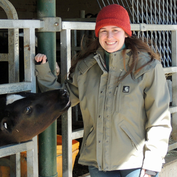 Nicole Welk-Joerger was photographed at a cow pen. She is wearing a red knit cap, and heavy gray jacket, and blue jeans. A cow is poking its head between the rails of the pen, reaching out to Welk-Joeger.