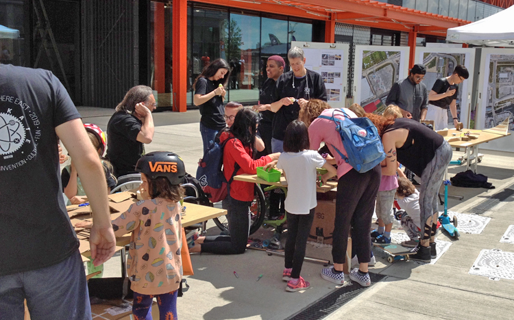 Groups of kids and their adults work at hands on projects at an outdoor table. One little girl is wearing a helmet emblazoned with the trademark “Vans.”