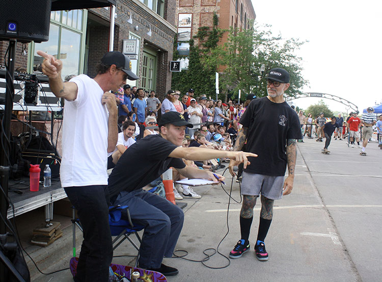 2 men pointing in different directions while a third looks on and a crowd in the background watches skaters perform tricks