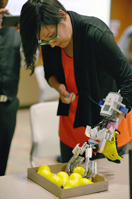 A woman bends over a box of yellow balls, trying to pick one up using the robotic hand that is attached to her arm.