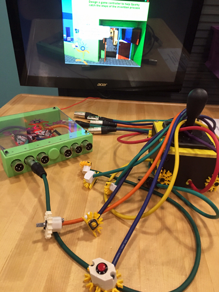 A tangle of purple, green, yellow, orange, and red cables with 3-prong connectors are arrayed on a table. A green cable is connected to a box of circuitry with a clear top and a green metal frame. Several cables wrap around a joystick; a red cable comes out of the joystick base. A computer screen at the end of the image displays a graphic of Sparky outside of SparkLab and the text “Design a game controller to help Sparky catch the steps of the invention process.”
