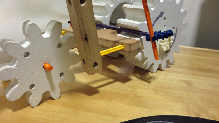 3 wooden gear-shaped wheels and wooden members with holes drilled in them are connected with Knex to form a 3-wheeled vehicle.