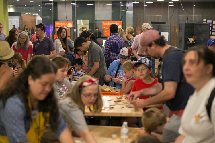 In the large main space of SparkLab, many, many children and adults are working on activities, talking, observing, and interacting with SparkLab staff. Two long tables covered with craft materials are visible, with children and their accompanying adults creating inventions. The photo was taken during the grand reopening of SparkLab in July 2015.
