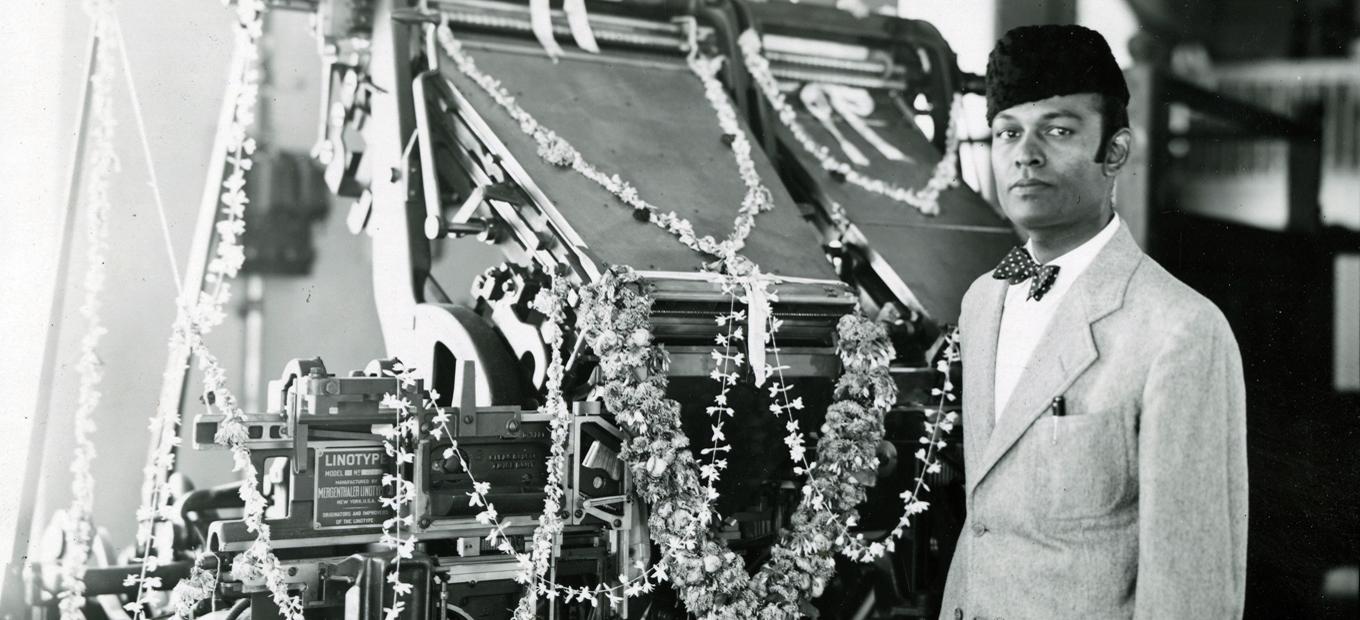 Hari Govil, wearing a sports jacket and bowtie, and a Karakul-type hat, stands in front to a Linotype machine festooned with flower garlands.