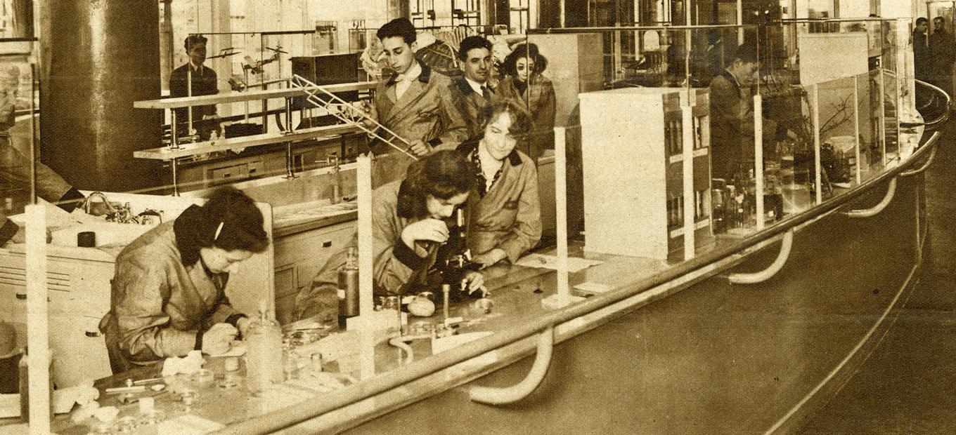A group of male and female students and faculty in lab coats at different stations in a science lab at the Westinghouse Fair World, 1940. Some are looking through microscopes, others at models of airplanes, still others at biological specimens.