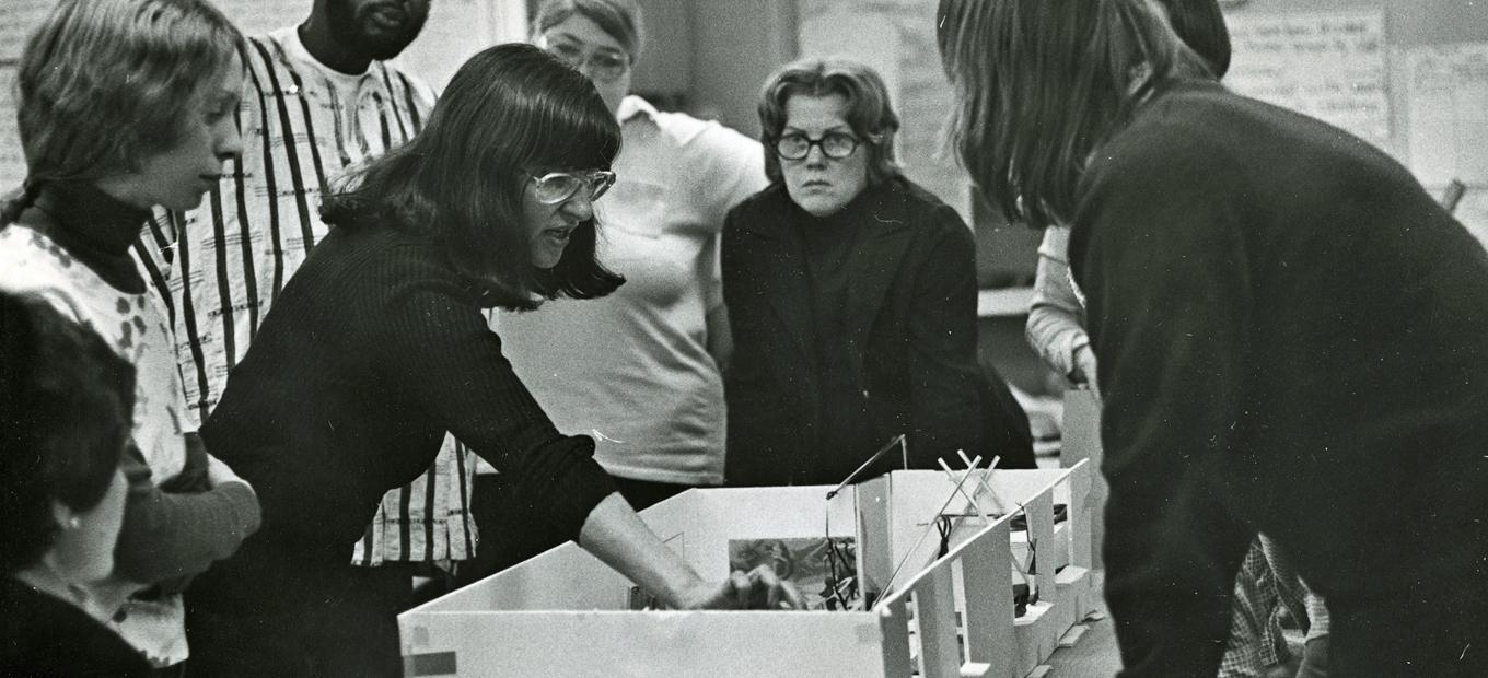 Elaine Ostroff, pointing at a model of a play room, with 6 staff members of Planning for Play around her, about 1975