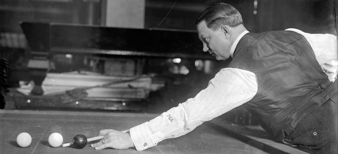 Side view of billiards great Ora C. Morningstar, bent over billiards table, cue stick in hand, about to take a shot.