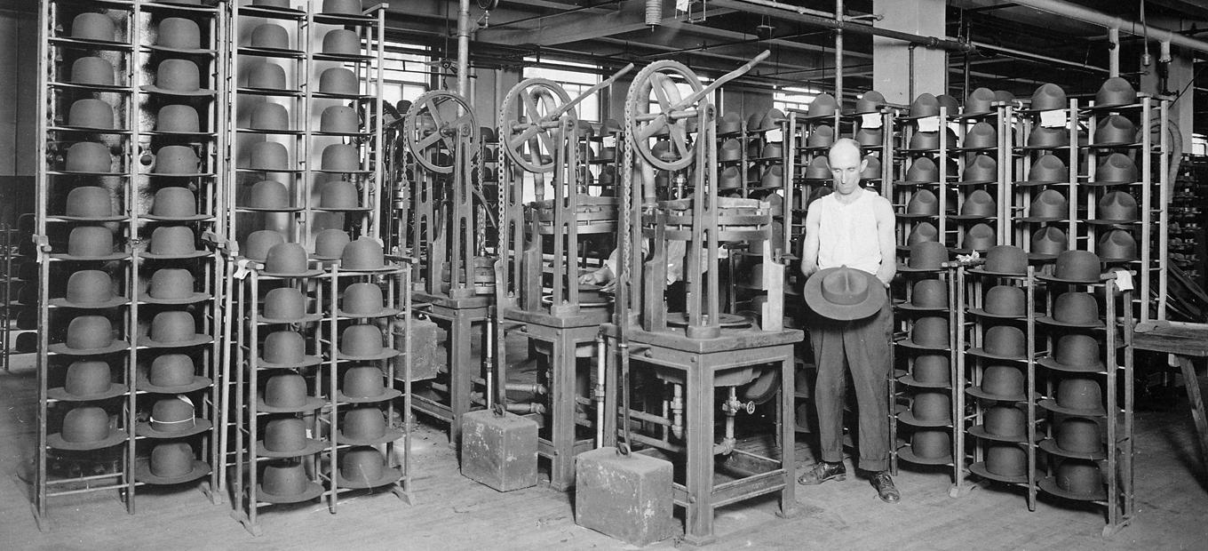 Worker standing next to a machine in the Stetson hat factory in Philadelphia, holding a hat and surrounded by racks of hats, around 1917