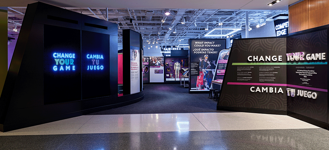 A head-on view of the entry way to the "Change Your Game/Cambia tu juego" exhibition. On the left is a video wall showing the exhibition logo, and on the right is the wall text with the exhibition title.