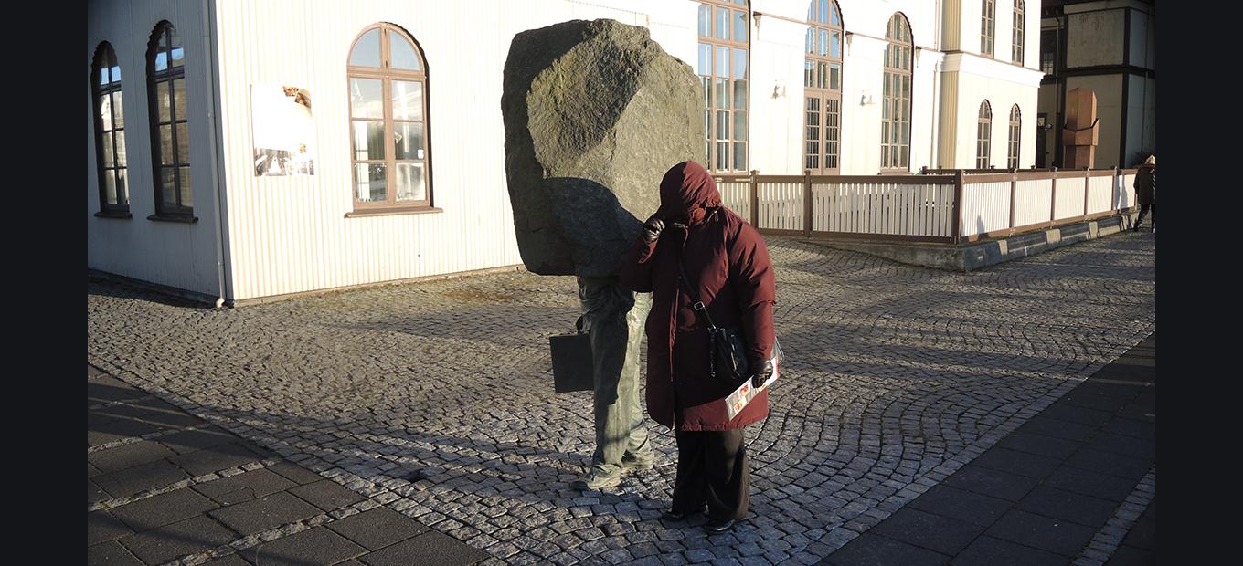 Woman in a winter coat standing next to a sculpture of a businessman whose head is a block of stone