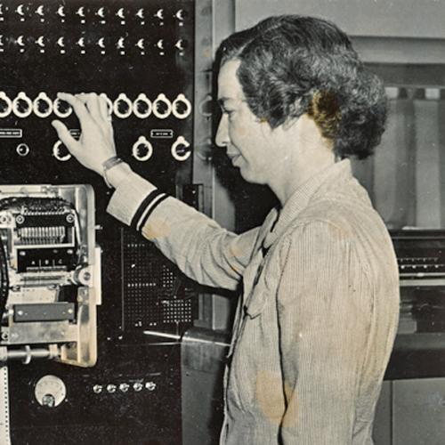 Grace Hopper, wearing her navy uniform, standing in profile to the camera. She has her right hand on one of the many dials that run across the top of the Mark I computer. She is looking at a section of the computer with many gears to move the punched tape code through the machine.