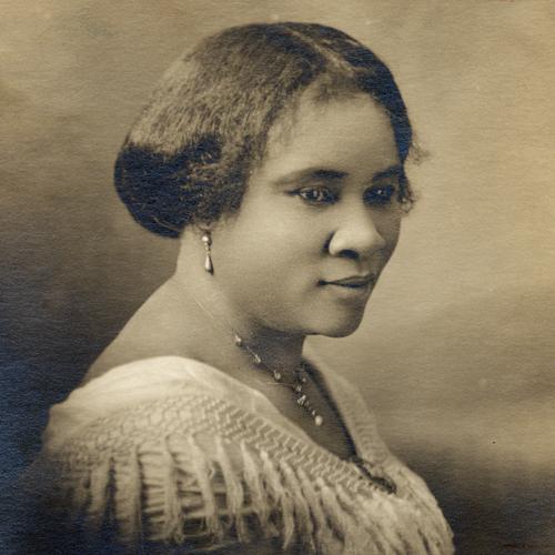 A sepia-toned 3/4 profile studio portrait photograph of Sarah Breedlove Walker, known as Madam C. J. Walker. She is wearing a shawl with tassels adorning the neckline, fastened with a brooch, and she wears drop earrings with matching necklace and a brooch.