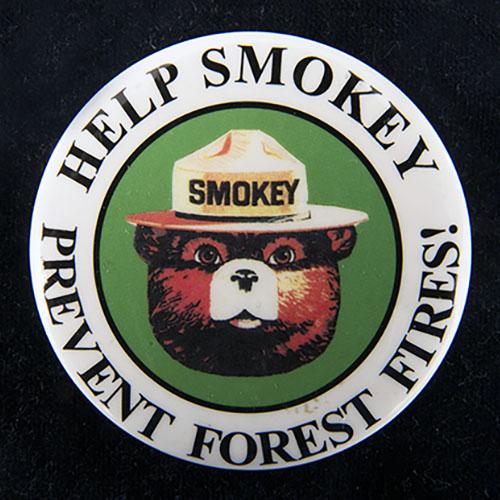 Metal button with image of Smokey the Bear’s head and the words “Help Smokey Prevent Forest Fires” around the outer edge