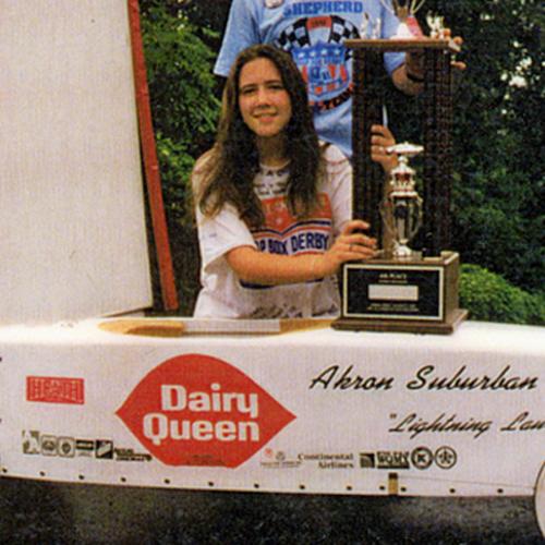 Detail of a photo of 14-year-old Laura Shepherd kneels behind her soapbox derby car, holding her trophy, which is resting on the car. 