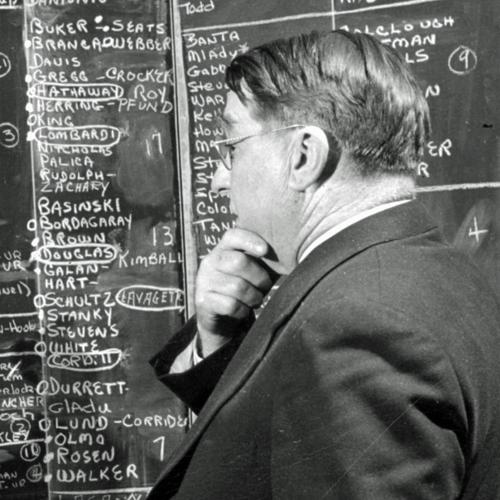 Branch Rickey holding his chin and looking at a blackboard with lists of players’ names