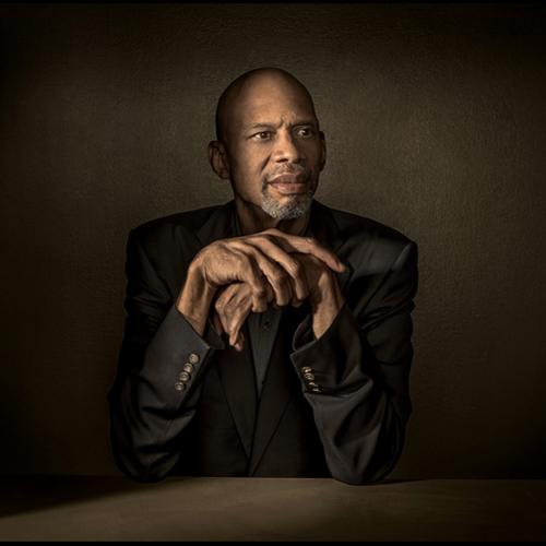 Kareem Abdul-Jabbar sitting at a table with the points of his elbows resting on the table and his hands loosely folded under his chin. He gazes off to the right. Dramatic lighting illuminates his face and hands; the rest of the image is in shadow.