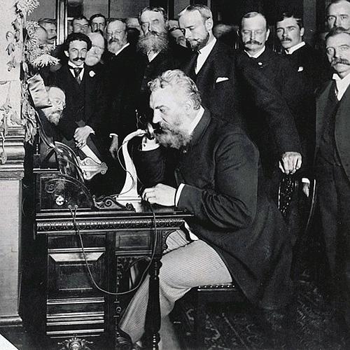 Alexander Graham Bell seated at table, speaking into telephone while a group of men watch.