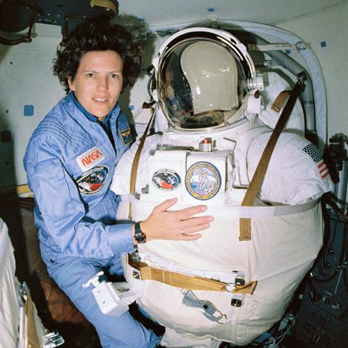 Kathryn D. Sullivan poses for a picture on 25 April 1990, before donning her spacesuit and extravehicular mobility unit in the airlock of the space shuttle Discovery. She is wearing a blue NASA jumpsuit and holding onto her inflated spacesuit.