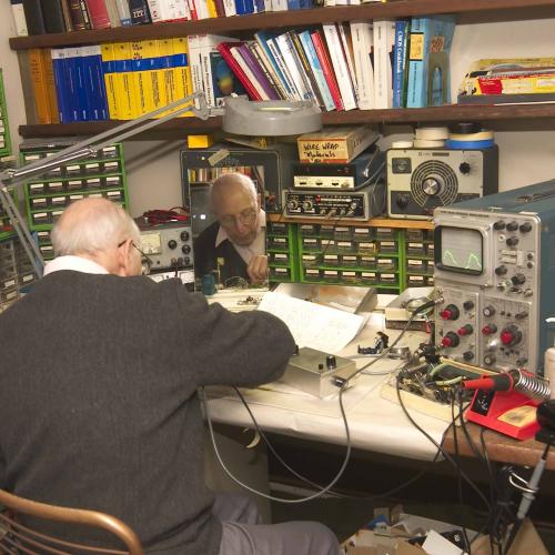 Ralph Baer is sitting at his desk, with his back to the camera, working on something. The desk is covered with equipment and parts and books cover the wall in front of him.
