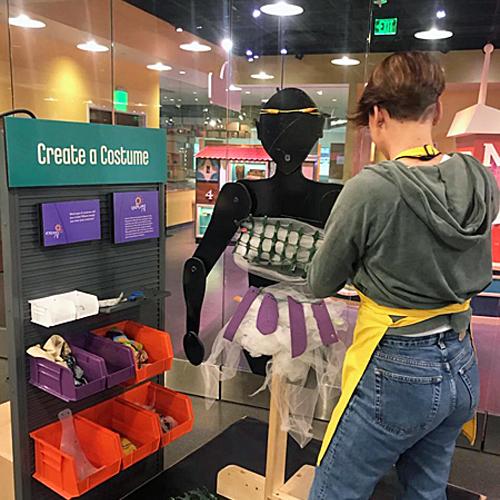 Hannah Correlli, a young woman with short, dark hair and wearing jeans, a hoodie, and a yellow SparkLab apron, stands with her back to the camera, working on an articulated human cutout form in the “Create a Costume” activity in SparkLab.