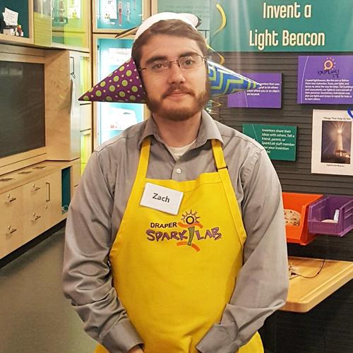 SparkLab facilitator Zach Etsch standing in SparkLab, wearing his yellow staff apron and a funny hat made from a propeller beanie and brightly-patterned paper cones over each ear.