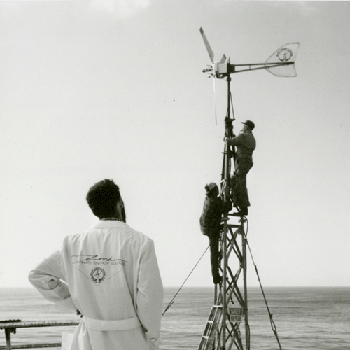Two workers stand near a wind turbine while a viewer in a lab coat observes from a distance.