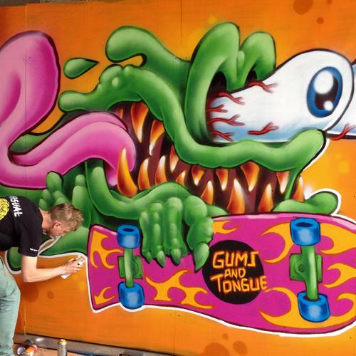 A graffiti artist is bent over, holding a spray paint can, finishing his colorful artwork that looks like a green slime monster with sharp teeth and bloodshot, bugged-out eyes, with its tongue flapping in the wind. The monster holds a skateboard reading, “Gums and Tongue.”